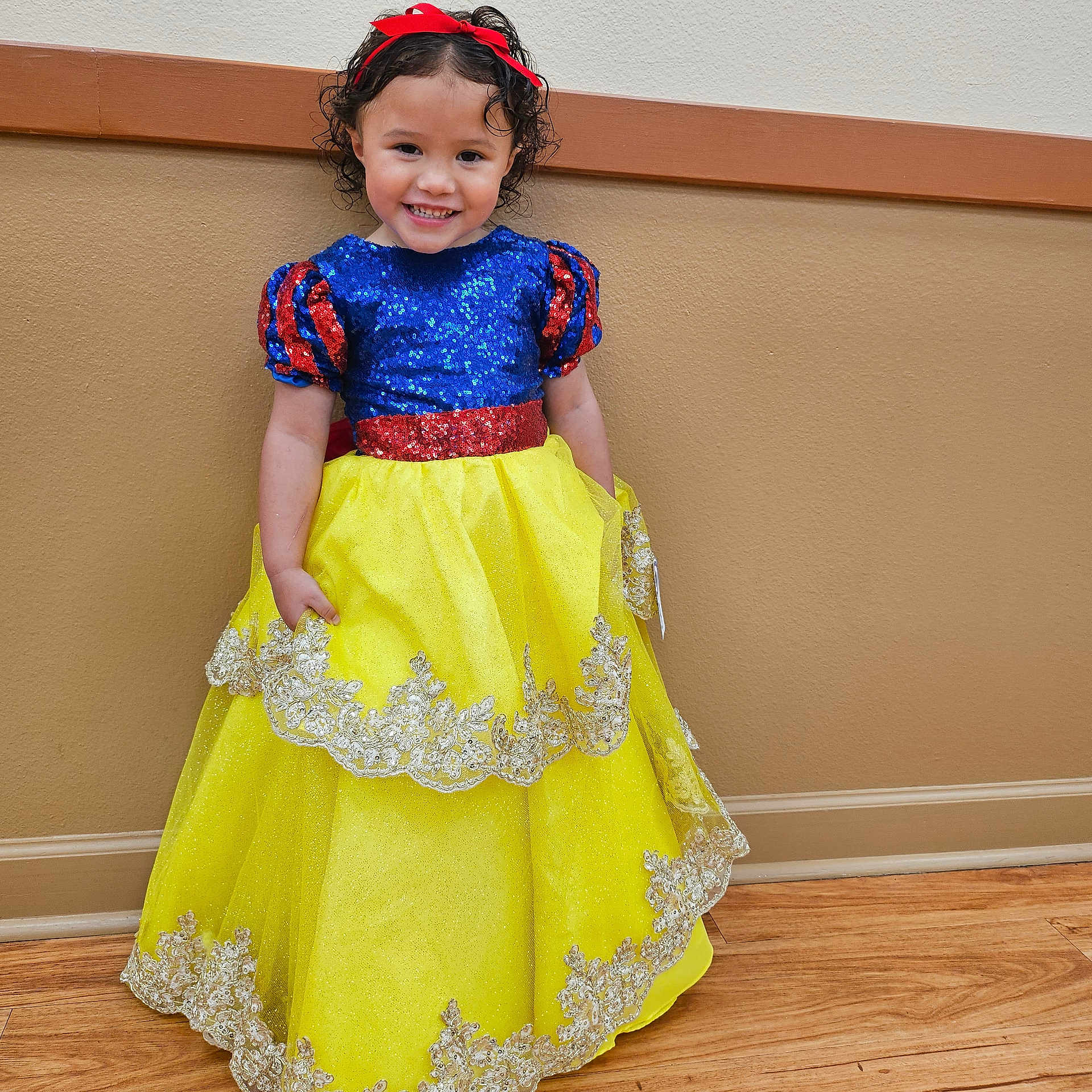 Ileanna is registered to the contest to win money with this photo: child, girl, smile, dress, costume, princess, sequins, yellow_skirt, blue_top, red_bow, curly_hair, indoor, wall, floor, happy, portrait, fashion, cute, standing, playful