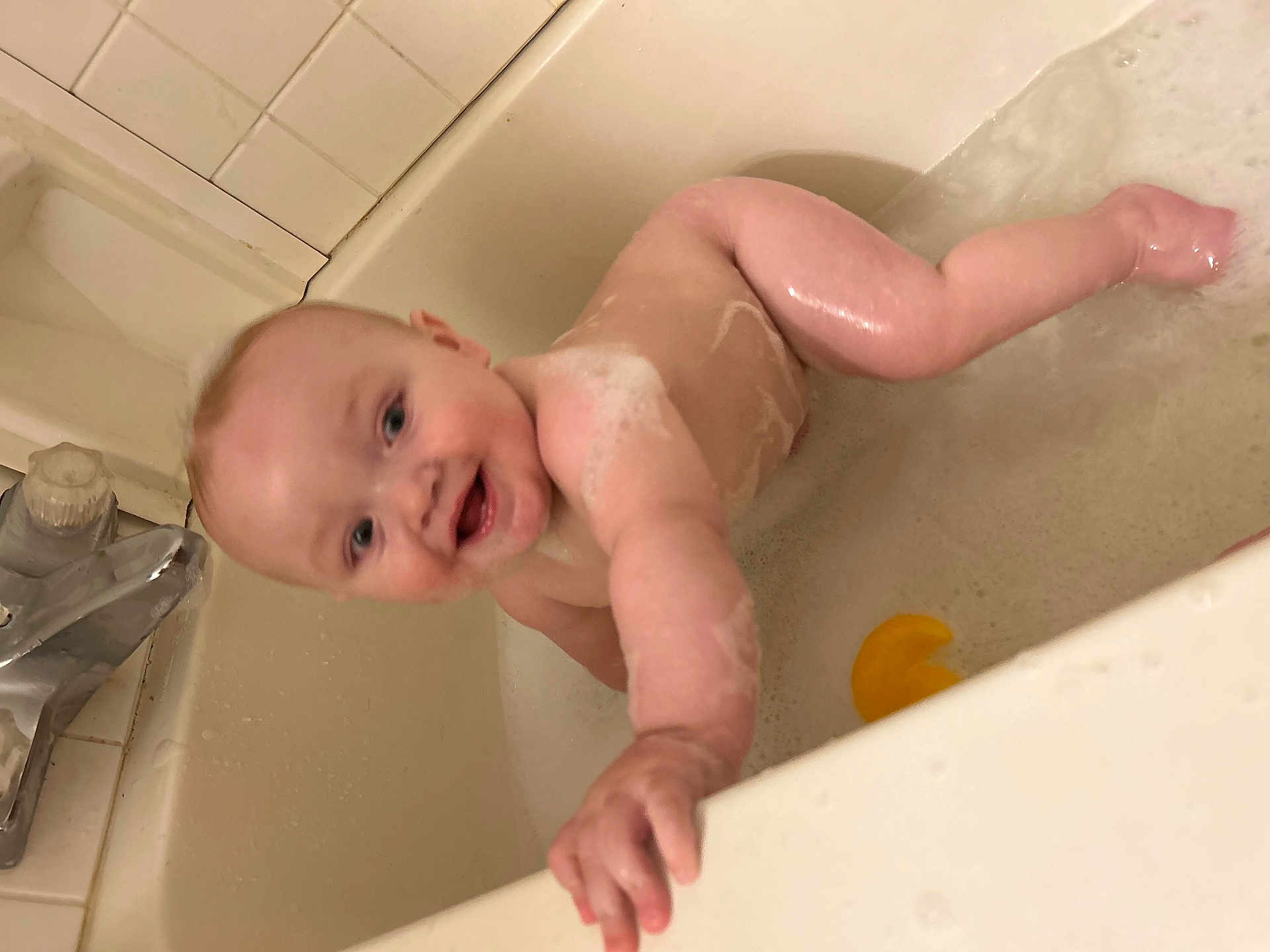 Elijah is registered to the contest to win money with this photo: baby, bath, bathtub, water, foam, rubber_duck, child, smile, skin, hand, face, happy, playing, splash, tiled_wall, faucet, soap_bubbles, bath_time, infant, cute