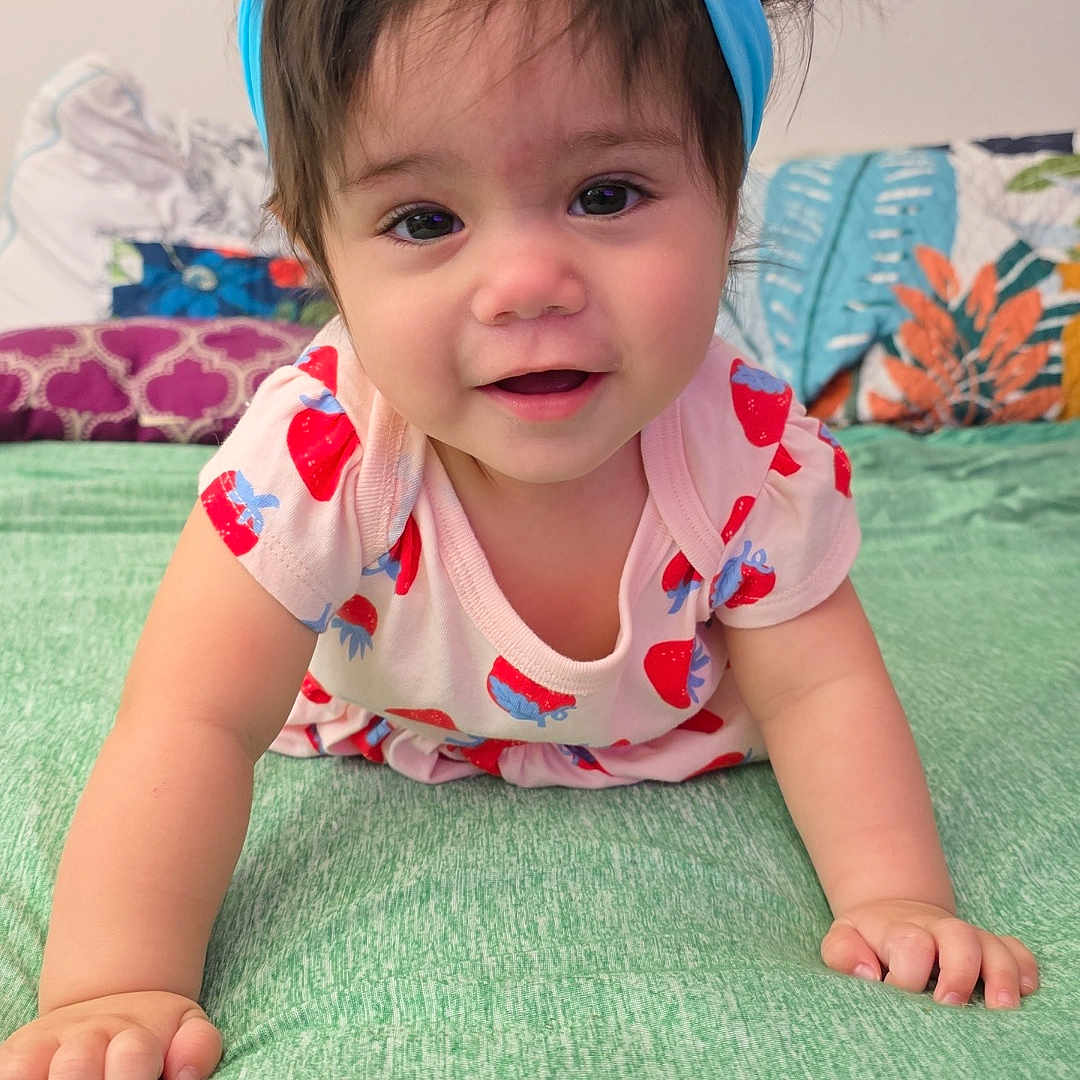 Elowyn joined the competition — help win amazing prizes! baby, child, crawling, headband, blue_bow, strawberry_pattern, pink_clothing, green_surface, pillow, colorful_background, indoors, cute, smiling, face, person, infant, young_child, happy, portrait, soft_lighting