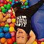 toddler, child, ball_pit, colorful_balls, laughing, upside_down, black_shirt, black_pants, indoor, play, fun, happy, smiling, cushion, plastic_balls, casual_clothing, young_child, joyful, play_area, active