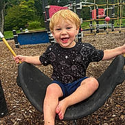 Waylon joined the competition — help win amazing prizes! child, toddler, smiling, happy, playground, swing, outdoor, park, blond_hair, barefoot, cosmic_shirt, black_shirt, shorts, sitting, laughing, play_equipment, rope_climbing, wood_chips, trees, portrait
