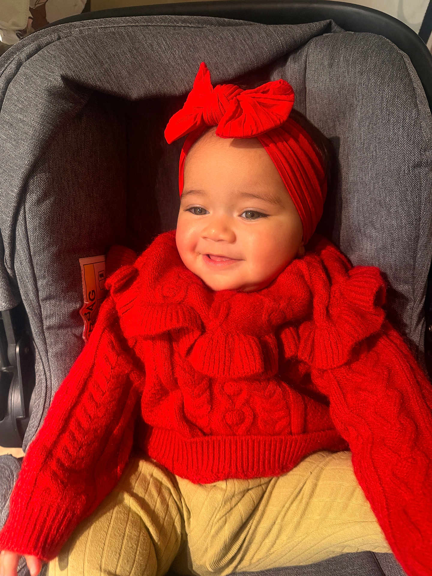 Prabhleen joined the competition — help win amazing prizes! baby, bow, child, cozy, cute, gray, happy, headband, indoors, infant, knitted_clothing, person, portrait, red_sweater, seated, smile, stroller, warm_lighting, yellow_pants, young_child