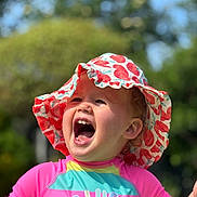 Arabella is registered to the contest to win money with this photo: child, toddler, hat, sunhat, laughing, smiling, outdoor, nature, colorful_clothing, rash_guard, summer, happy, portrait, closeup, sunlight, greenery, face, headwear, playful, daylight