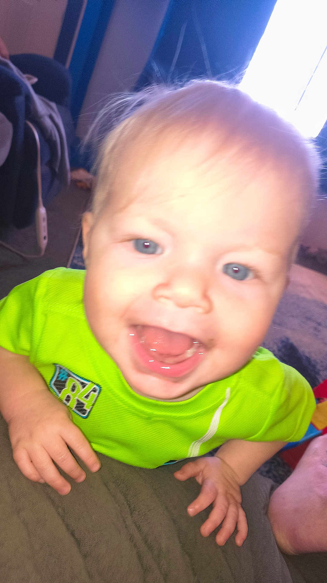 Lyall joined the competition — help win amazing prizes! baby, infant, child, smiling, open_mouth, blue_eyes, blond_hair, green_shirt, close_up, hands, tongue, teeth, drool, indoor, couch, blanket, window_light, playful, portrait, overexposed