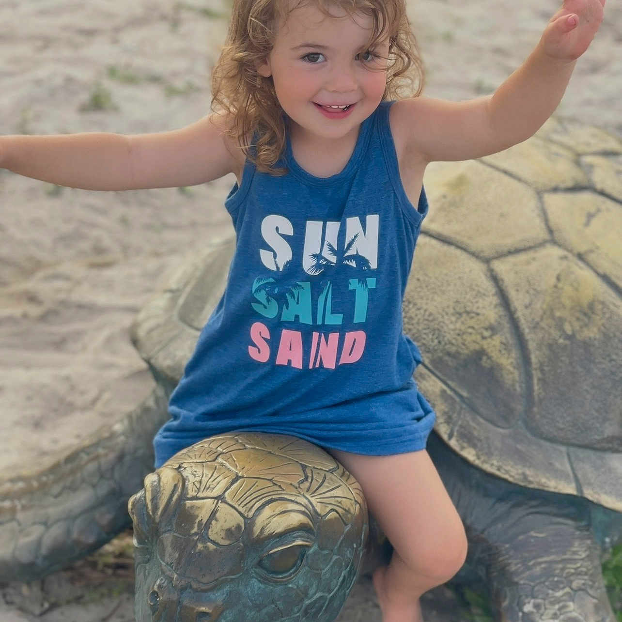 Adalynn is registered to the contest to win money with this photo: child, toddler, smiling, outdoor, beach, sand, turtle, turtle_sculpture, statue, blue_shirt, summer, portrait, arms_raised, sitting, barefoot, playful, curly_hair, animal, travel, daylight