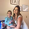 child, girl, adult, woman, smiling, portrait, family, indoor, kitchen, table, snack_bag, wall_sign, calendar, tattoo, dress, blue_shirt, brown_hair, seated, happy, photograph