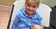 Adalynn joined the competition — help win amazing prizes! black_shorts, blonde_hair, blue_jacket, bulletin_board, casual_clothing, chair, child, door, filing_cabinet, girl, happy, indoors, office, photograph, portrait, seated, smiling, staff_shoutout, tile_floor, toddler