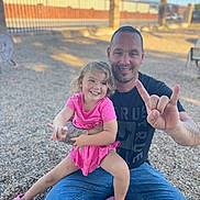 Adalynn is registered to the contest to win money with this photo: child, girl, father, family, smiling, happy, outdoor, portrait, sitting, lap, pink_dress, pink_shoes, tattoos, hand_gesture, gravel, fence, blue_sky, sunlight, playground, family_photo
