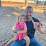 child, girl, father, family, smiling, happy, outdoor, portrait, sitting, lap, pink_dress, pink_shoes, tattoos, hand_gesture, gravel, fence, blue_sky, sunlight, playground, family_photo