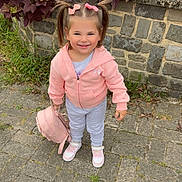 Lenaya participe au concours pour gagner de l'argent avec cette photo : child, toddler, girl, pigtails, pink_jacket, hair_bows, backpack, sneakers, smile, stone_wall, plants, sidewalk, outdoors, cute, looking_at_camera, full_body, standing, pavement, greenery, casual_clothing