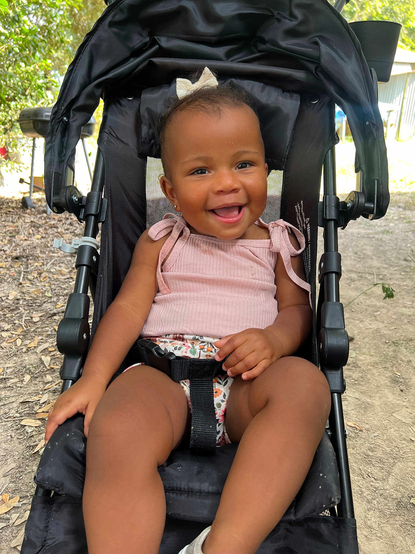Jocelyn is registered to the contest to win money with this photo: toddler, child, stroller, smiling, outdoor, nature, baby, happy, black_stroller, pink_top, bow, seatbelt, sunlight, leaves, ground, portrait, sitting, cute, person, daylight