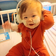 Logan joined the competition — help win amazing prizes! baby, child, hospital, bed, medical, nasal_tubes, headphones, orange_outfit, smiling, happy, indoor, furniture, person, healthcare, cute, infant, medical_tape, hospital_room, baby_girl, sitting