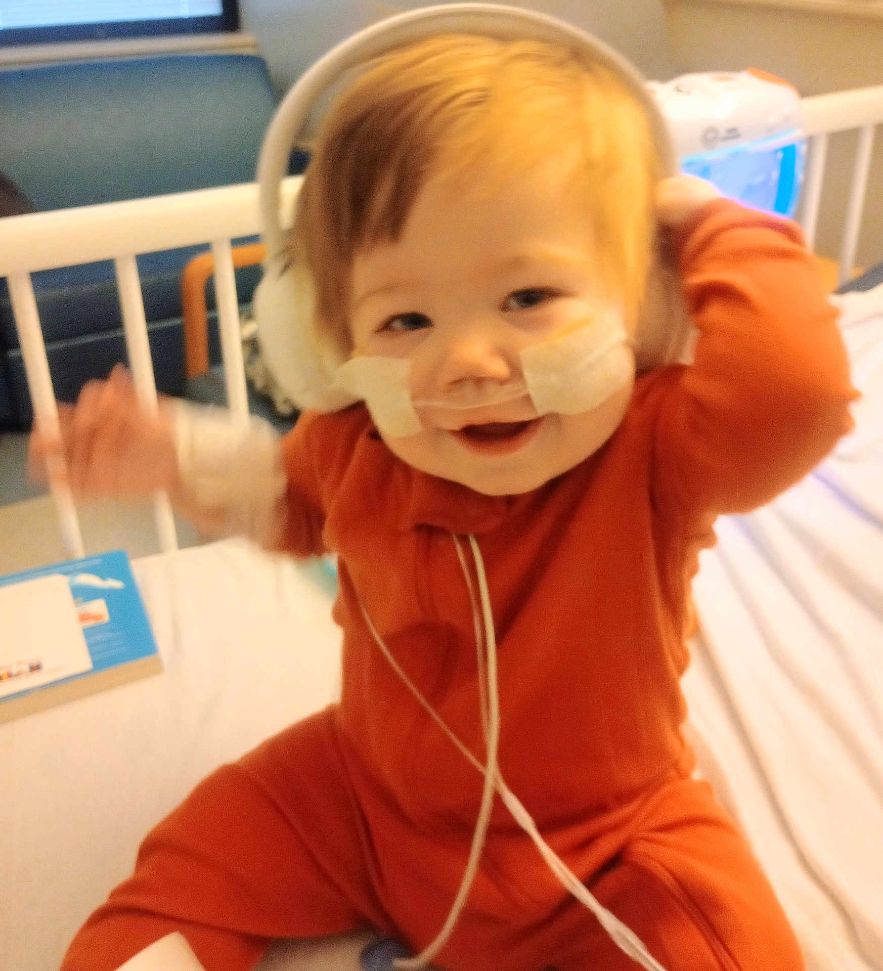 Logan joined the competition — help win amazing prizes! baby, child, hospital, bed, medical, nasal_tubes, headphones, orange_outfit, smiling, happy, indoor, furniture, person, healthcare, cute, infant, medical_tape, hospital_room, baby_girl, sitting