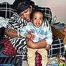 Giovanni joined the competition — help win amazing prizes! adult, baby, child, smile, indoor, person, seated, couch, patterned_clothing, blanket, headwear, toy, water_bottle, footwear, carpet, table, happy, holding, black_hair, tattoo