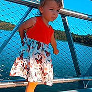 Mylana a rejoint le concours — aidez-le/la à gagner de superbes lots ! child, toddler, girl, dress, butterfly_print, orange_top, sandals, bridge, railing, metal, chainlink_fence, lake, water, sky, outdoors, wooden_deck, portrait, standing, pony_tail, sunny_day