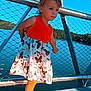 child, toddler, girl, dress, butterfly_print, orange_top, sandals, bridge, railing, metal, chainlink_fence, lake, water, sky, outdoors, wooden_deck, portrait, standing, pony_tail, sunny_day