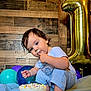 toddler, cake, birthday, balloons, number_one, frosting, celebration, wooden_wall, blue_striped_overalls, white_shirt, barefoot, party, colorful, cute, child, food, smudged_face, festive, indoor, squatting