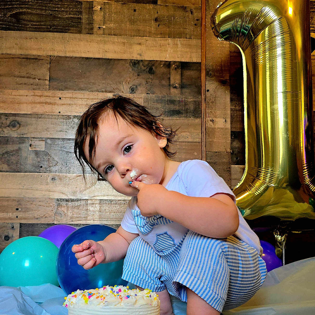 Oliver is registered to the contest to win money with this photo: balloons, barefoot, birthday, blue_striped_overalls, cake, celebration, child, colorful, cute, festive, food, frosting, indoor, number_one, party, smudged_face, squatting, toddler, white_shirt, wooden_wall