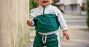 Ayub a rejoint le concours — aidez-le/la à gagner de superbes lots ! toddler, child, walking, tracksuit, green_clothing, white_clothing, sneakers, sidewalk, urban, city, building, wall, outdoor, daylight, person, young_child, curly_hair, smiling, standing, street
