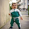 toddler, child, walking, tracksuit, green_clothing, white_clothing, sneakers, sidewalk, urban, city, building, wall, outdoor, daylight, person, young_child, curly_hair, smiling, standing, street