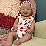 baby, infant, smiling, sitting, headband, strawberry_pattern, clothing, blanket, couch, happy, cute, indoor, portrait, child, furniture, skin, feet, hand, face, yellow_necklace