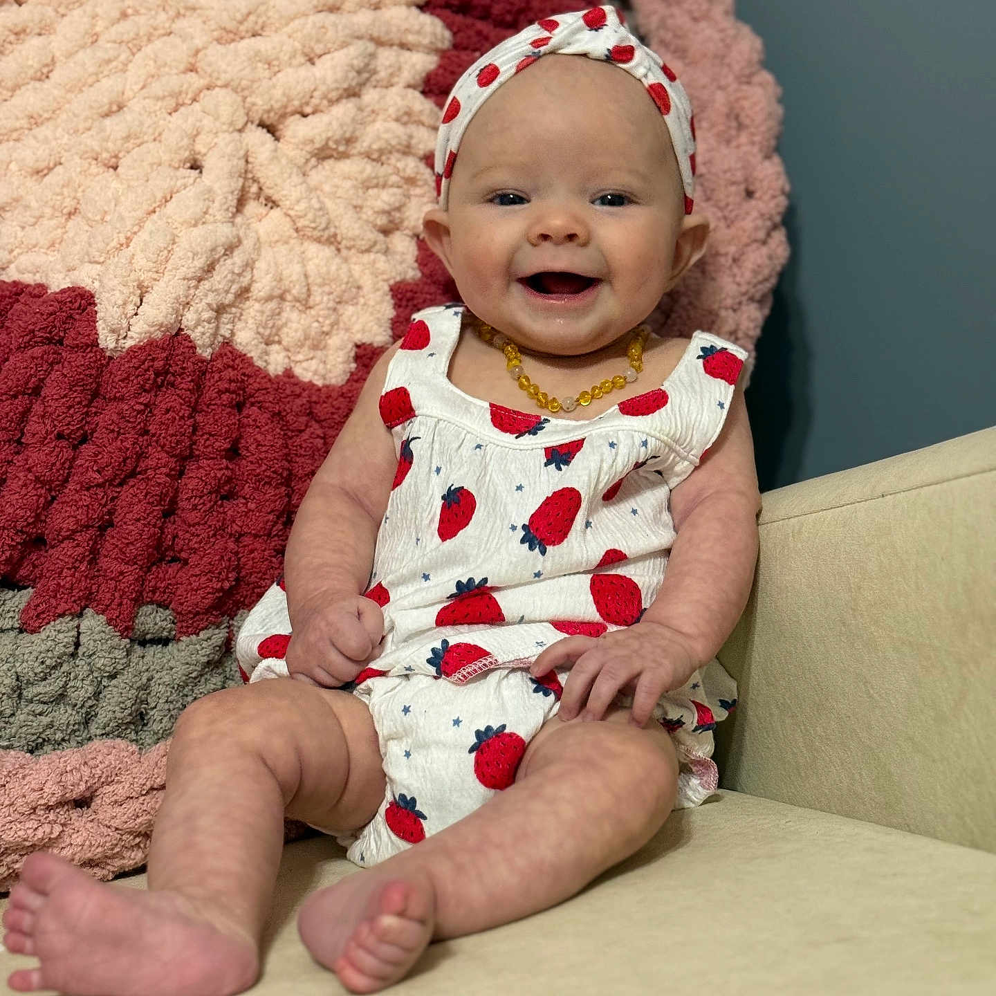 Emma Kaye is registered to the contest to win money with this photo: baby, blanket, child, clothing, couch, cute, face, feet, furniture, hand, happy, headband, indoor, infant, portrait, sitting, skin, smiling, strawberry_pattern, yellow_necklace