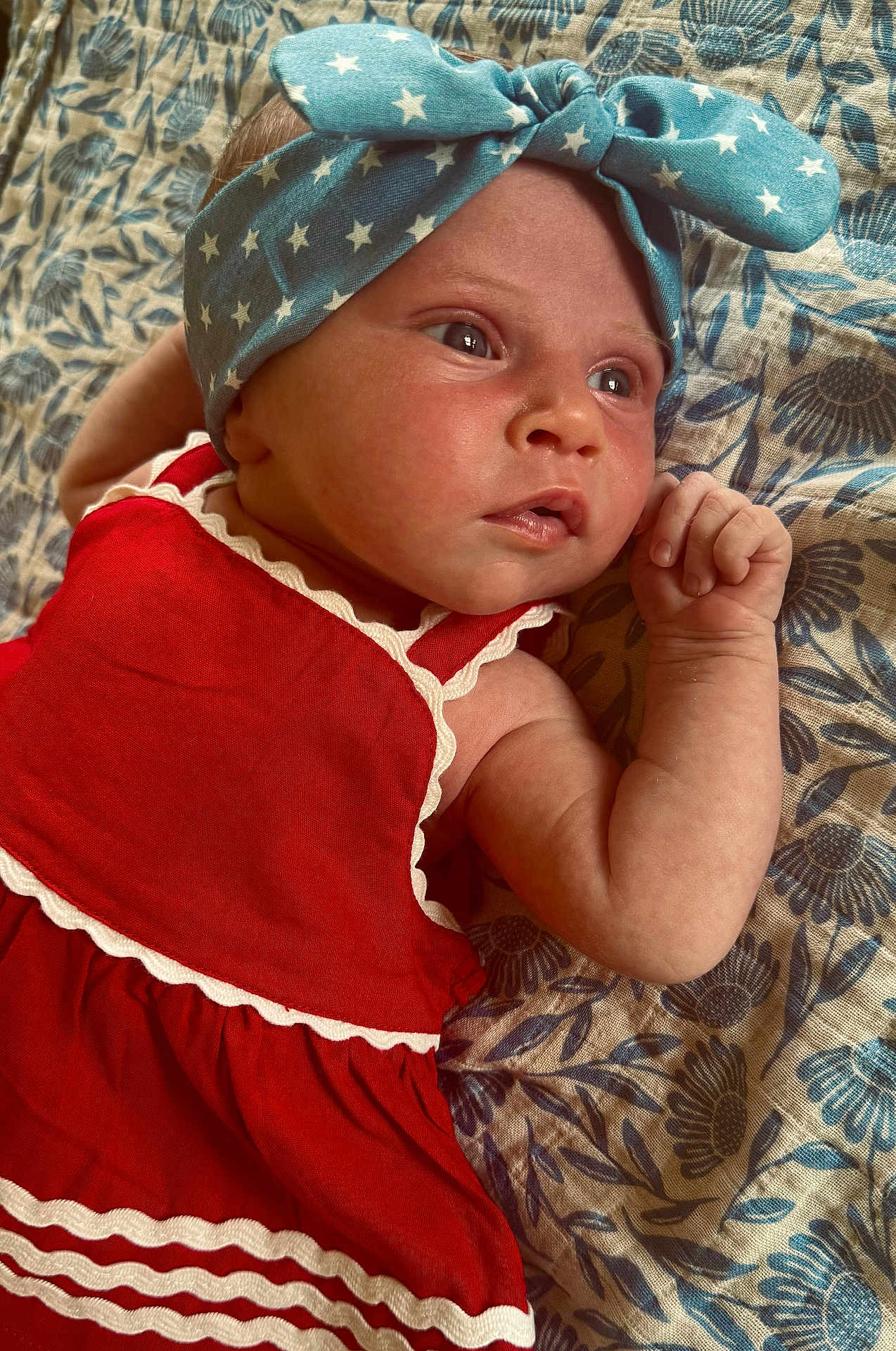 Delaney is registered to the contest to win money with this photo: baby, child, infant, headband, red_dress, blue_headband, stars, blanket, floral_pattern, skin, face, hand, lying_down, closeup, portrait, cute, indoors, expression, young, person