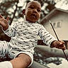 baby, child, infant, portrait, outdoor, sitting, pajamas, mustache_pattern, necklace, stick, hand, smiling, looking_at_camera, low_angle, trees, house, background, reflection, daylight, photography