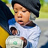 baby, infant, child, portrait, beanie, black_hat, white_clothing, dollar_bill, money, currency, hand, sleepy, outdoor, sunlight, close_up, face, person, carried, green_background, humorous