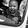 Da'Vonnie joined the competition — help win amazing prizes! baby, child, smiling, happy, portrait, black_and_white, monochrome, close_up, face, teeth, indoor, toy_car, steering_wheel, dashboard, playtime, sitting, hands, sweater, infant, photography
