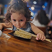Aria is registered to the contest to win money with this photo: child, curly_hair, noodles, chopsticks, wooden_table, restaurant, food, eating, cute, portrait, indoors, bokeh, face, person, meal, dining, casual, young, appetite, focus