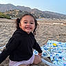 toddler, child, smiling, beach, sand, blanket, curly_hair, black_hoodie, shorts, outdoor, nature, mountains, evening, sky, happy, person, playful, relaxing, cute, landscape