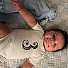 baby, infant, onesie, mustache_design, blanket, stuffed_animal, blue, white_clothing, smiling, lying_down, cute, child, soft_toy, indoor, portrait, happy, toddler, face, hand, cozy