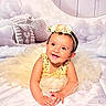 baby, bed, bedding, birthday_style, child, cloud_background, face, flower_crown, gold_dress, hands, happy, indoor, infant, pillow, portrait, sequins, sitting, smiling, studio_photo, stuffed_animal