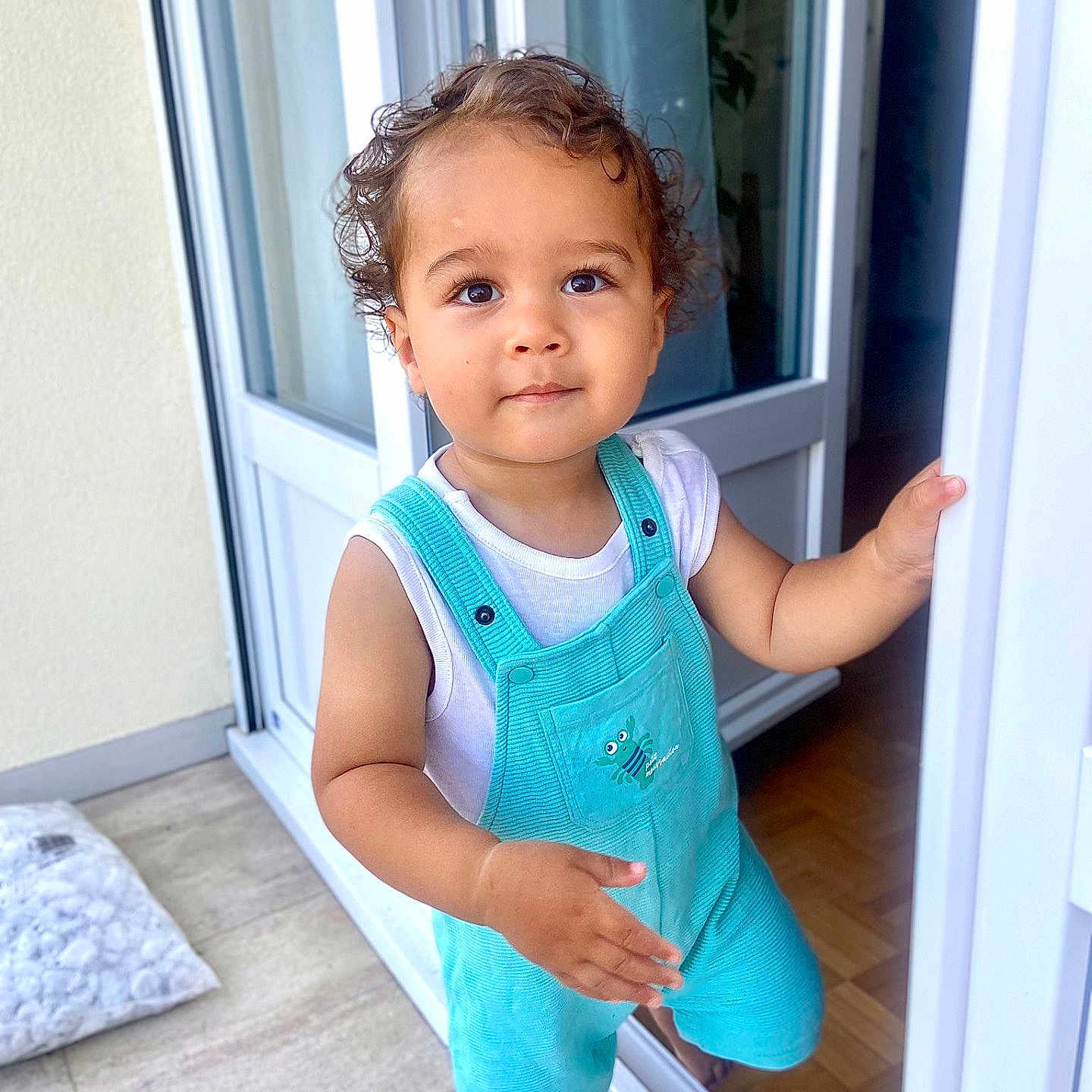 Andréa a rejoint le concours — aidez-le/la à gagner de superbes lots ! baby, child, curly_hair, curtain, cute, doorway, floor, hand, home, indoors, looking_up, outdoors, overalls, person, portrait, standing, toddler, turquoise, white_shirt, window