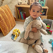 Andréa participe au concours pour gagner de l'argent avec cette photo : toddler, child, plush_toy, bunny, carpet, cushion, teddy_bear, books, toy_blocks, shelf, sock, yellow_towel, indoor, smiling, hair_tie, wooden_furniture, soft_toy, playroom, baby, floor