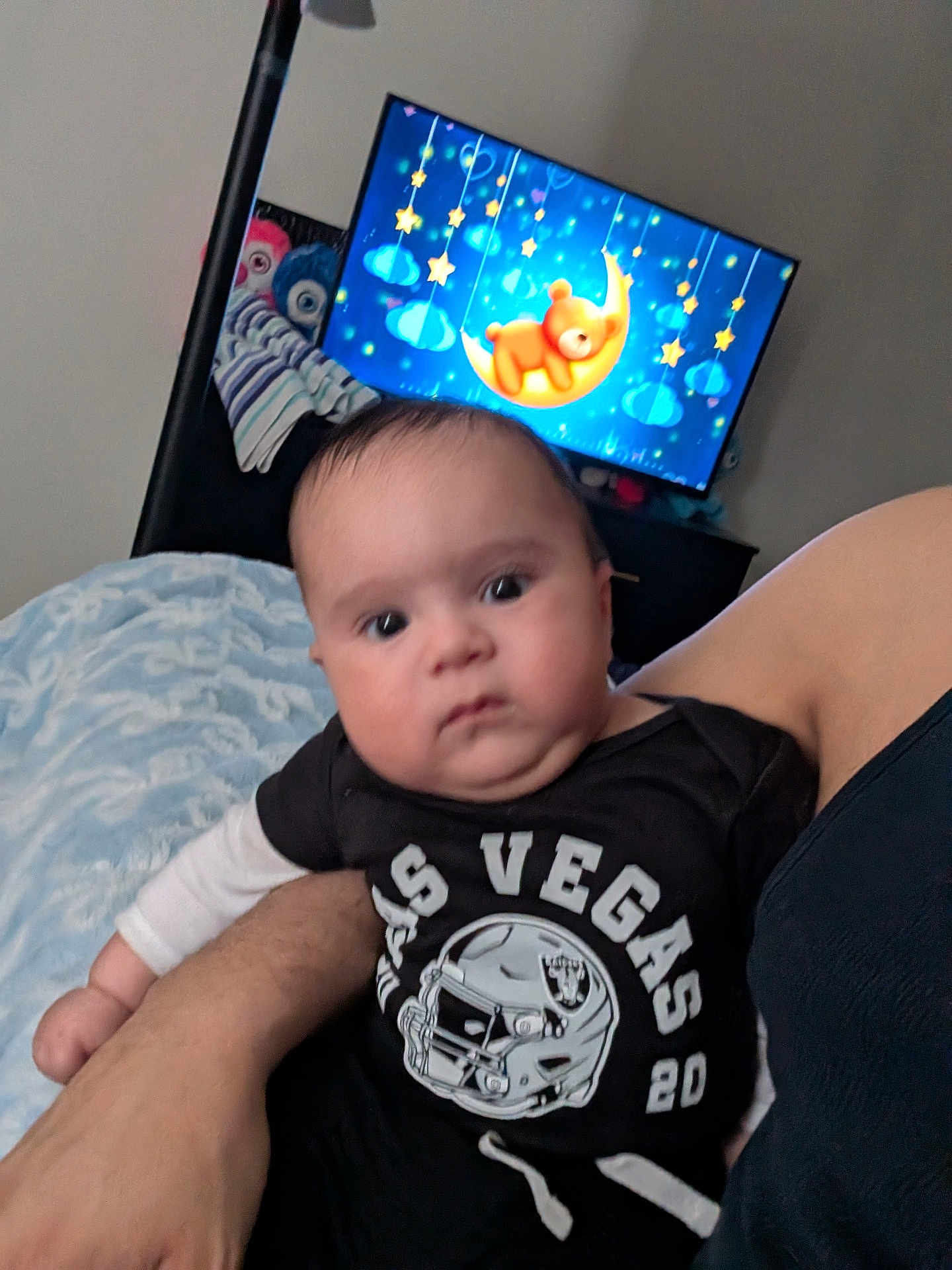 Joe is registered to the contest to win money with this photo: baby, infant, child, television, cartoon, moon, teddy_bear, bed, blanket, arm, shirt, las_vegas, sports_logo, portrait, indoors, room, staring, cute, holding, parent