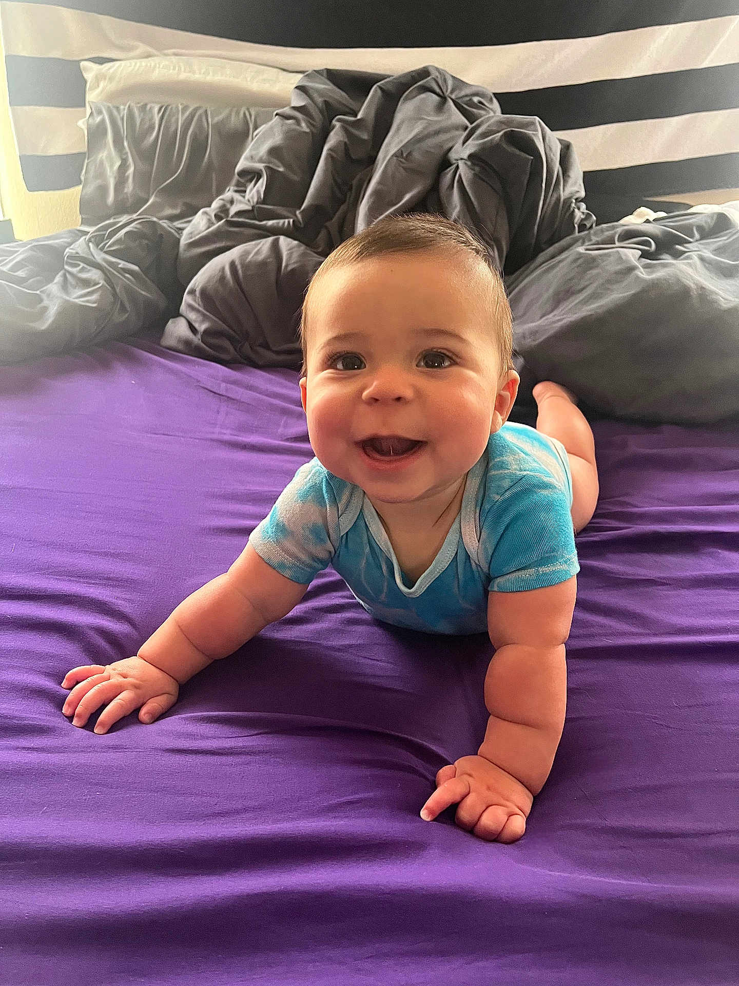 Isaiah joined the competition — help win amazing prizes! baby, infant, smiling, blue_clothing, purple_bedspread, bed, blanket, pillow, indoor, child, cute, happy, toddler, person, face, arms, skin, head, eyes, mouth