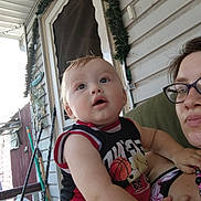 Robert is registered to the contest to win money with this photo: baby, child, woman, glasses, porch, door, garland, outdoor, fence, curious, expression, holding, basketball_shirt, person, face, arm, hand, clothing, beige_skin, head