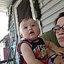 baby, child, woman, glasses, porch, door, garland, outdoor, fence, curious, expression, holding, basketball_shirt, person, face, arm, hand, clothing, beige_skin, head