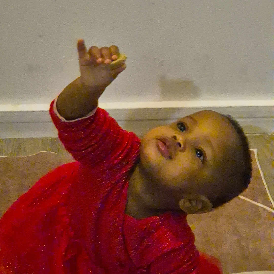 Fatoumata a rejoint le concours — aidez-le/la à gagner de superbes lots ! baby, carpet, child, curious, cute, expression, floor, indoor, kneeling, long_sleeves, looking_up, person, playful, portrait, reaching, red_dress, small_hand, toddler, wall, young_child