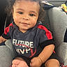 toddler, child, car_seat, curly_hair, smiling, casual_clothing, shirt, text_on_clothing, indoor, car_interior, seatbelt, foot, red_black_clothing, young_child, portrait, cute, happy, person, baby, young_kid