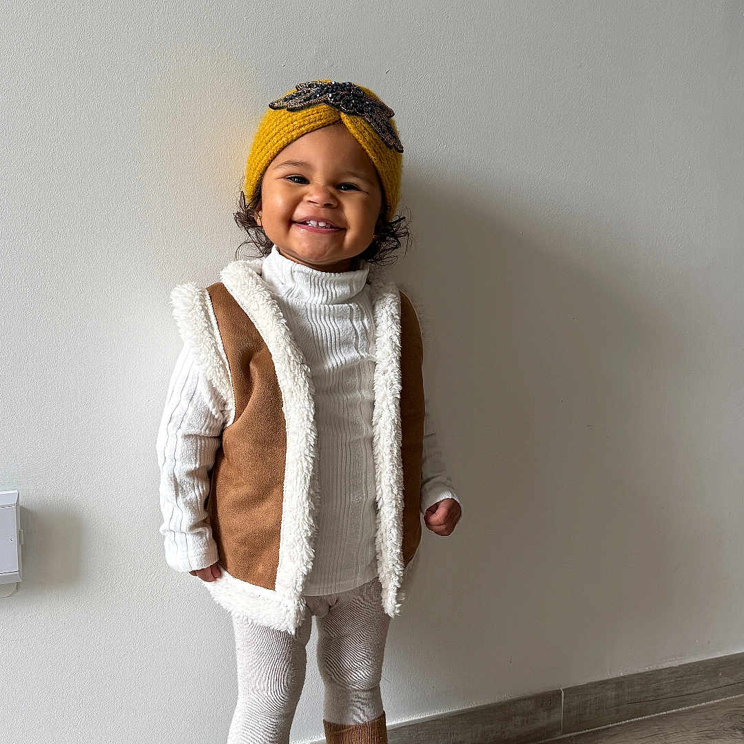 Lyria a rejoint le concours — aidez-le/la à gagner de superbes lots ! black_boots, brown_vest, child, cozy_outfit, curly_hair, cute, happy, indoor, knit_headband, leggings, minimal_wall, one_person, portrait, shearling, smiling, standing, toddler, white_turtleneck, wood_floor, yellow_hat
