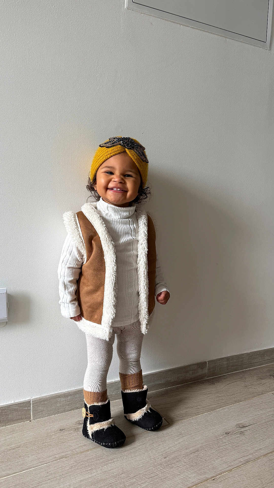 Lyria a rejoint le concours — aidez-le/la à gagner de superbes lots ! child, toddler, smiling, yellow_hat, knit_headband, curly_hair, white_turtleneck, brown_vest, shearling, leggings, black_boots, standing, indoor, minimal_wall, wood_floor, portrait, cute, cozy_outfit, happy, one_person