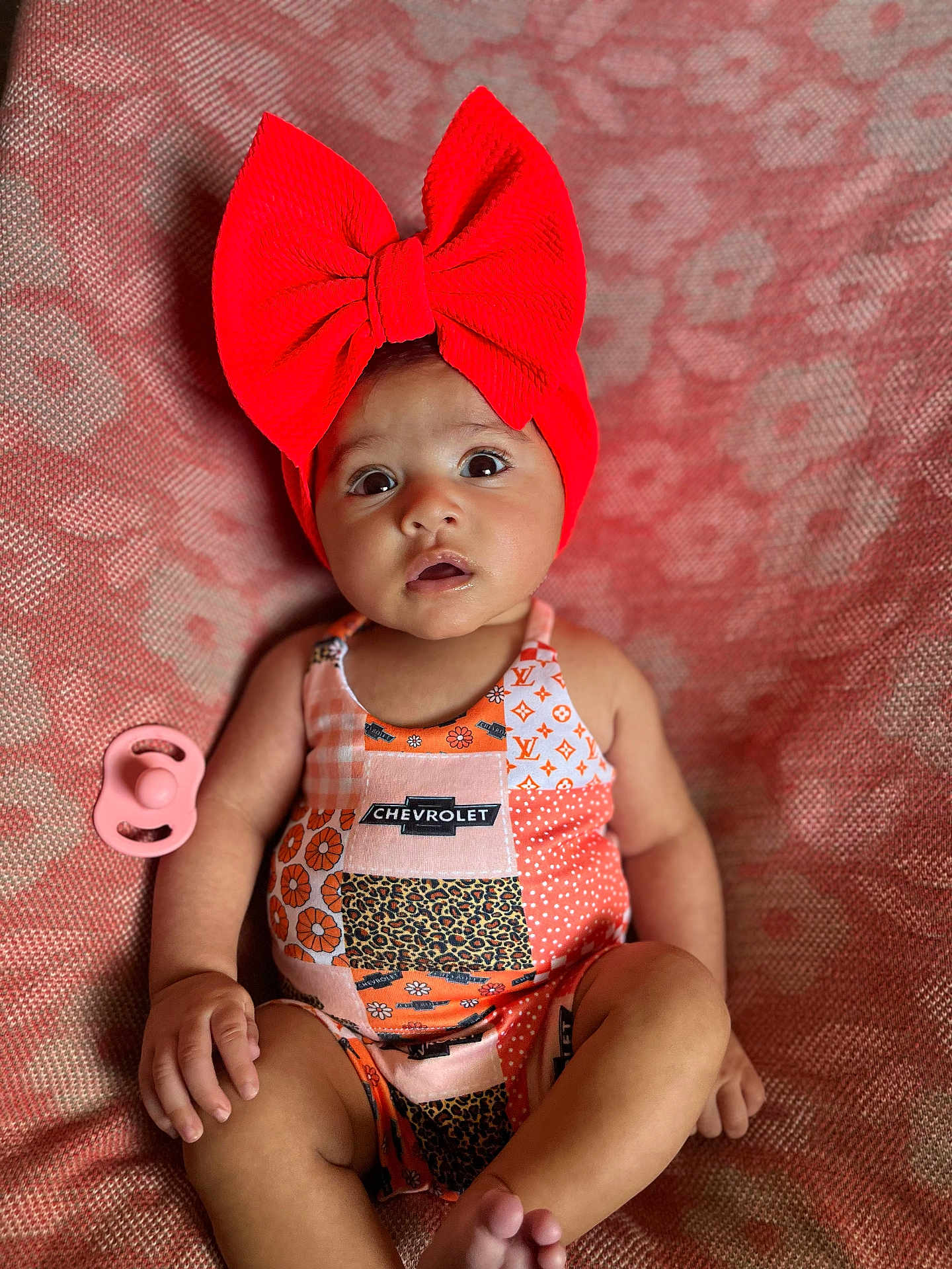 Oaklynn is registered to the contest to win money with this photo: baby, infant, red_bow, headband, chevrolet_logo, colorful_outfit, patchwork, pacifier, blanket, patterned_blanket, sitting, wide_eyes, cute, child, indoors, portrait, skin, hand, foot, expression