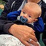 baby, pacifier, blanket, bear_pattern, adult_hand, ring, jacket, outdoor, bench, chain_link_fence, cloudy_sky, person, child, blue_eyes, face, holding, casual_clothing, closeup, portrait, comfort