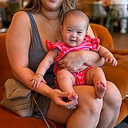 Aria is registered to the contest to win money with this photo: baby, woman, smile, indoor, couch, orange, pink_clothing, person, holding, happy, arm, face, legs, handbag, casual, shorts, shoes, background, blurred, seated