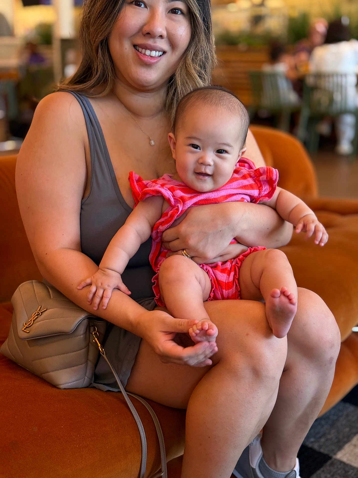 Aria is registered to the contest to win money with this photo: baby, woman, smile, indoor, couch, orange, pink_clothing, person, holding, happy, arm, face, legs, handbag, casual, shorts, shoes, background, blurred, seated