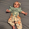 baby, child, infant, portrait, lying_down, bed, bedspread, floral_outfit, dress, headband, bow, wide_eyed, looking_at_camera, arms, hands, barefoot, indoor, home, gray_background, fashion