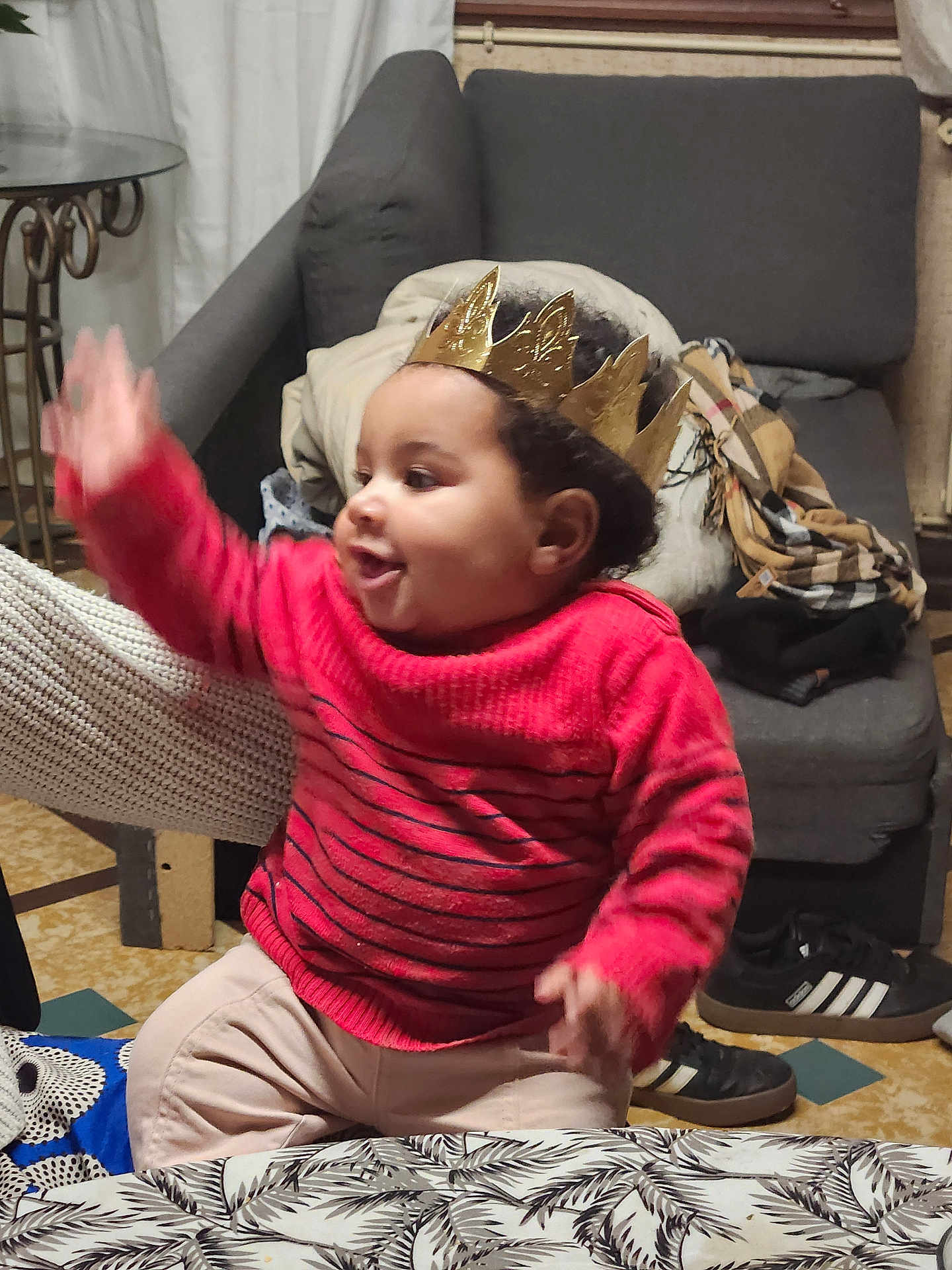 Eva a rejoint le concours — aidez-le/la à gagner de superbes lots ! toddler, child, crown, red_sweater, indoor, sofa, table, shoes, curtain, blanket, cheerful, smiling, person, floor, pattern, furniture, home, curly_hair, seated, gesture